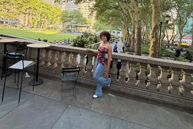 A young woman poses beside a railing, with a tree-lined park and buildings behind her.
